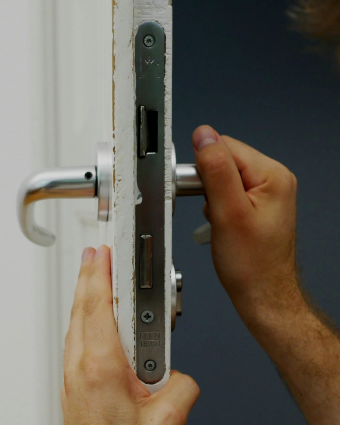 Two hands fitting a mortise lock and a lever handle into the edge of a white door